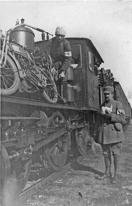Junan kyydissä seisoo mies moottoripyörän kanssa ja takana ohjaamon ikkunasta kurkistaa toinen mies. Junan vieressä maassa seisoo kolmas mies.